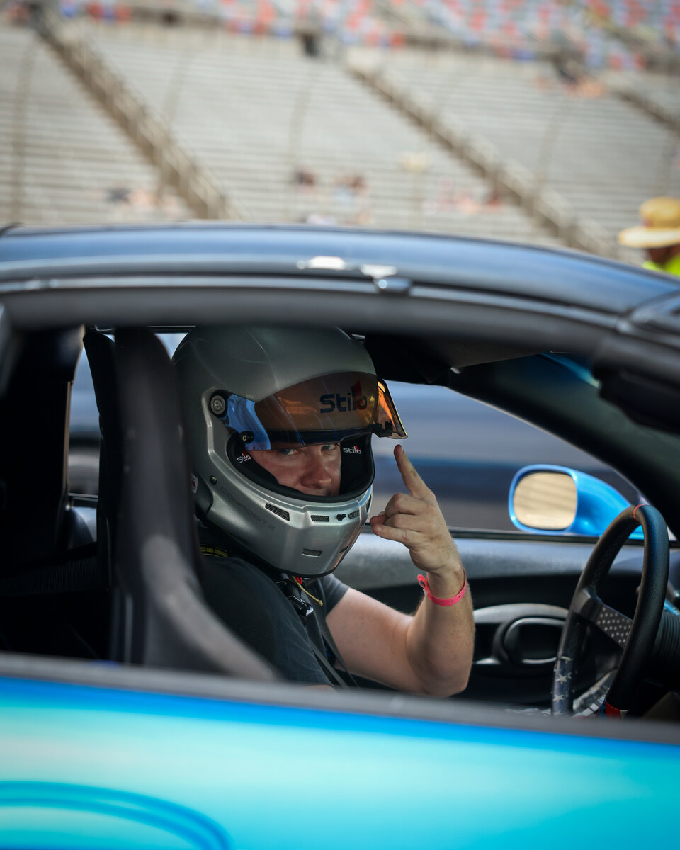 Driver seated in a race car wearing a white helmet, hands on the wheel, ready for a session