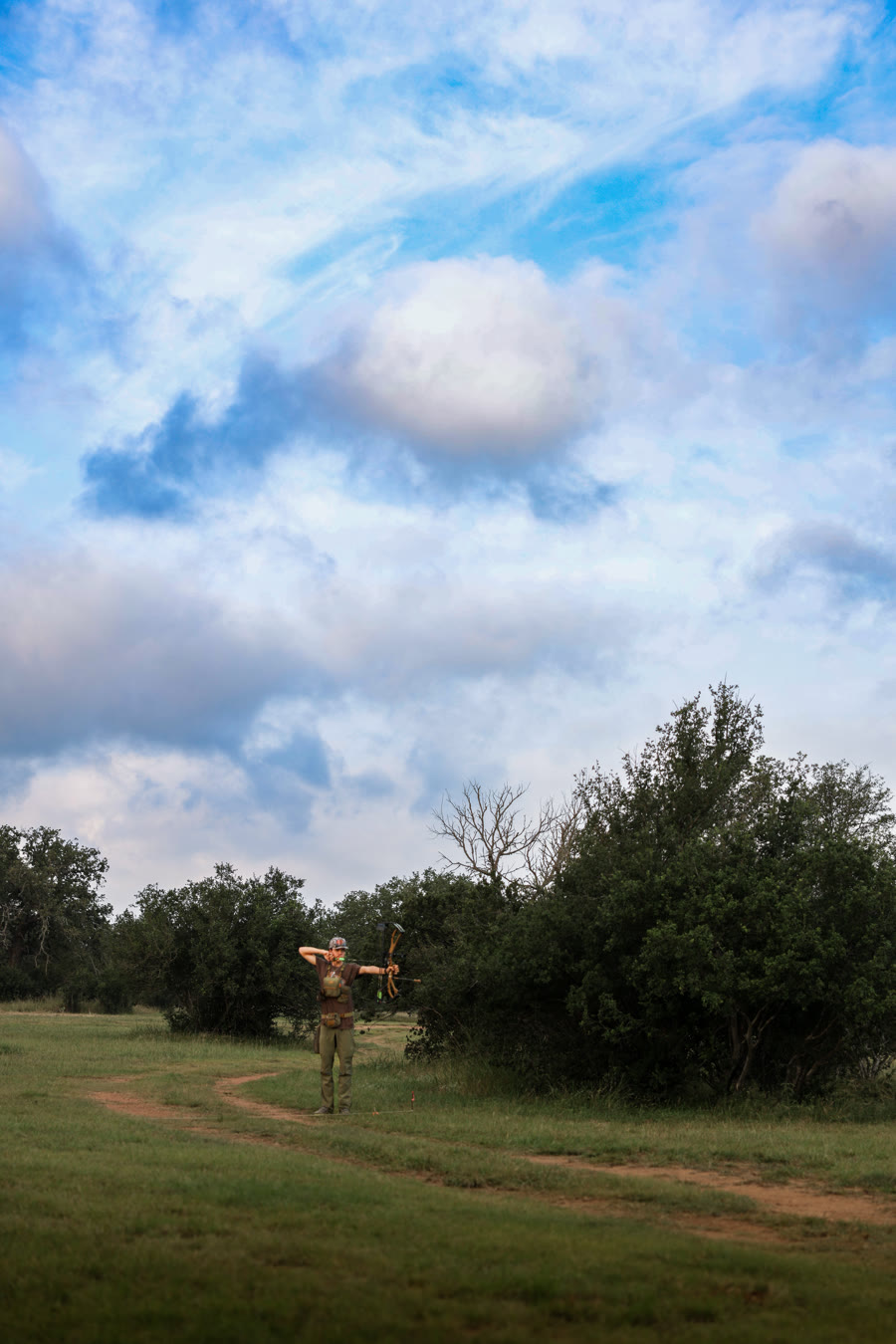 Archer at full draw on a dirt path under a dramatic cloudy sky